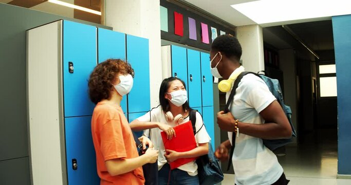 Group Of 3 Teenage Multiethnic Students Wearing Medical Masks Greet Each Other And Talk In The Hallway Of The School