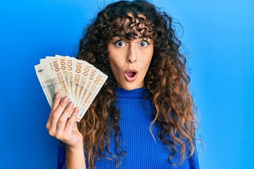 Young hispanic girl holding 100 danish krone banknotes scared and amazed with open mouth for...