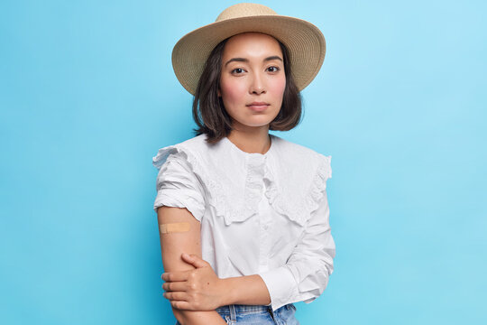Serious beautiful dark haired Asian woman wears sunhat and white blouse shows arm with plaser after successful covid vaccination models against blue background. Coronavirus immunization concept
