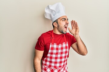 Young hispanic man wearing baker uniform shouting and screaming loud to side with hand on mouth. communication concept.