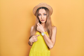 Young blonde girl wearing summer hat in hurry pointing to watch time, impatience, looking at the...