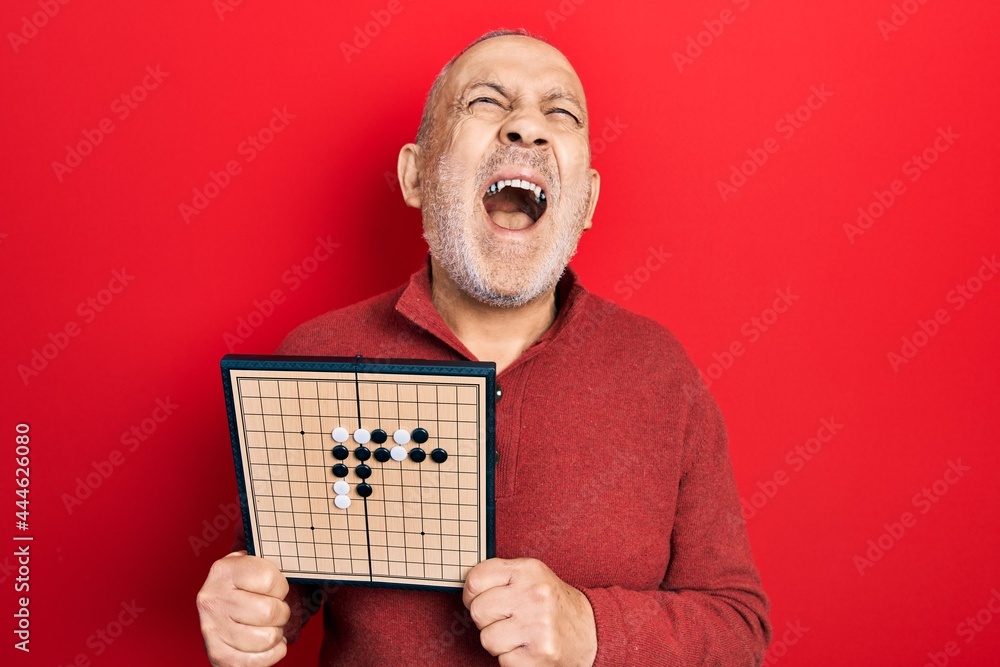 Sticker Handsome mature man holding asian go game board angry and mad screaming frustrated and furious, shouting with anger looking up.