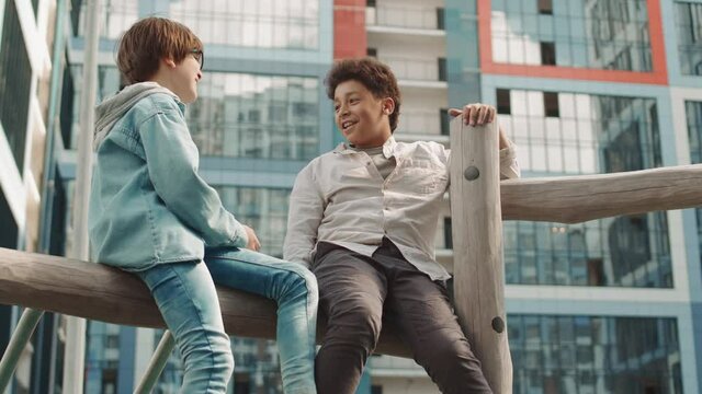 Slowmo shot of multiethnic schoolboys sitting on top of climber playset at playground in schoolyard having conversation