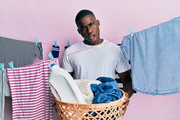 Young african american man holding laundry basket in shock face, looking skeptical and sarcastic,...