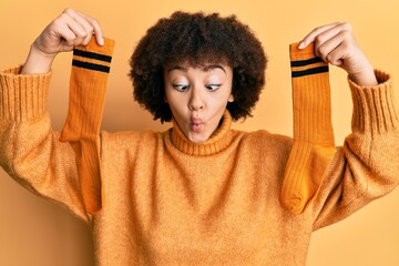 Young hispanic girl holding pair of socks making fish face with mouth and squinting eyes, crazy and...