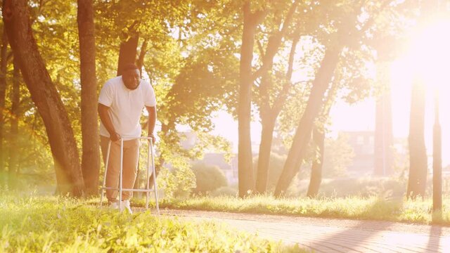 Disabled African-American man is walking with a walker. Handicapped patient in the park. Assistance, rehabilitation and health care. Golden hour sunset.