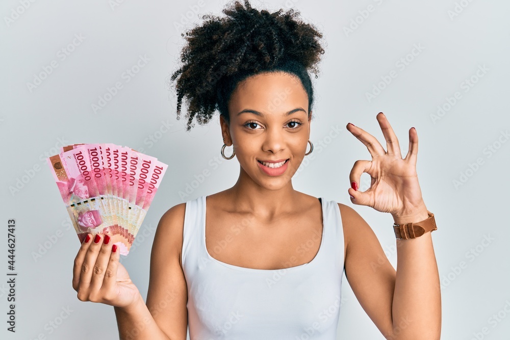 Poster Young african american girl holding indonesian rupiah banknotes doing ok sign with fingers, smiling friendly gesturing excellent symbol