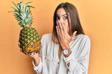 Young beautiful woman holding pineapple covering mouth with hand, shocked and afraid for mistake....