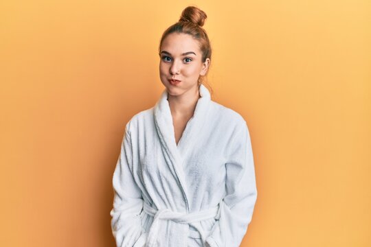 Young blonde woman wearing bathrobe puffing cheeks with funny face. mouth inflated with air, crazy expression.