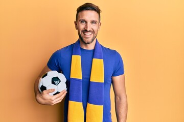 Handsome man with beard football hooligan cheering game holding ball looking positive and happy standing and smiling with a confident smile showing teeth