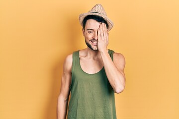 Young hispanic man wearing summer hat covering one eye with hand, confident smile on face and surprise emotion.