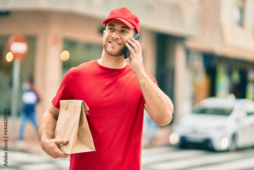 Sticker Young caucasian deliveryman talking on the smartphone and holding delivery paper bag at the city.