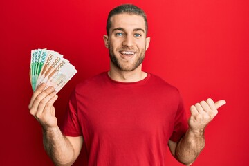 Young caucasian man holding 5000 south korean won banknotes pointing thumb up to the side smiling happy with open mouth