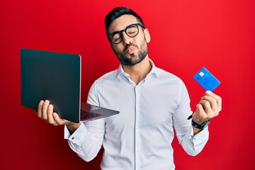 Young hispanic man wearing business style holding laptop and credit card looking at the camera...