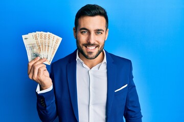 Young hispanic businessman wearing business suit holding denmark krone banknotes looking positive and happy standing and smiling with a confident smile showing teeth
