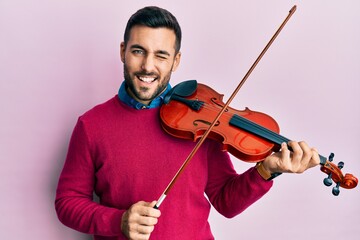 Young hispanic musician man playing violin winking looking at the camera with sexy expression,...