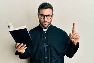 Young hispanic priest man holding bible with finger up skeptic and nervous, frowning upset because...