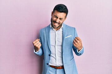 Young hispanic businessman wearing business jacket excited for success with arms raised and eyes...