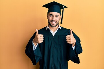 Young hispanic man wearing graduation cap and ceremony robe success sign doing positive gesture...