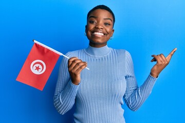 Young african american woman holding tunisia flag smiling happy pointing with hand and finger to the side