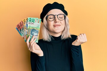 Young blonde girl holding australian dollars screaming proud, celebrating victory and success very excited with raised arm