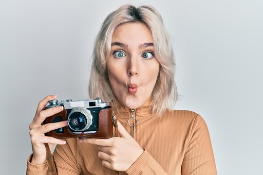 Young blonde girl holding vintage camera making fish face with mouth and squinting eyes, crazy and comical.