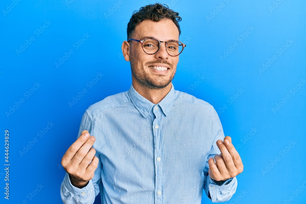 Wall mural young hispanic man wearing casual clothes and glasses doing money gesture with hands, asking for sal