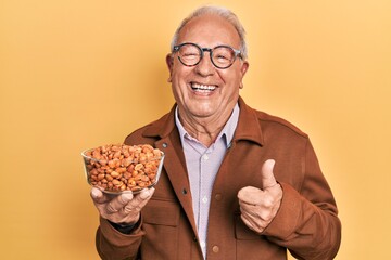 Senior man with grey hair holding fried peanuts smiling happy and positive, thumb up doing excellent and approval sign