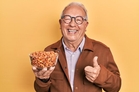 Senior man with grey hair holding fried peanuts smiling happy and positive, thumb up doing excellent and approval sign