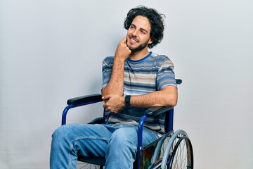 Handsome hispanic man sitting on wheelchair with hand on chin thinking about question, pensive...
