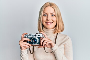 Young blonde woman holding vintage camera looking positive and happy standing and smiling with a confident smile showing teeth - Powered by Adobe