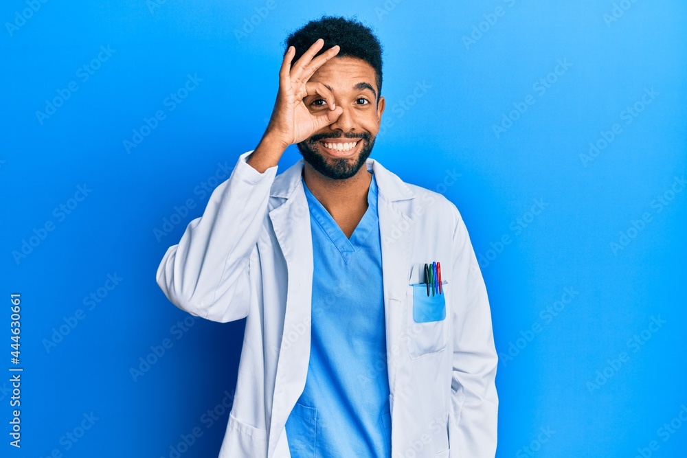 Wall mural Handsome hispanic man with beard wearing doctor uniform doing ok gesture with hand smiling, eye looking through fingers with happy face.