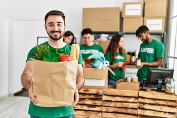 Group of young volunteers working at charity center. Man smiling happy and holding paper bag with food to donate.