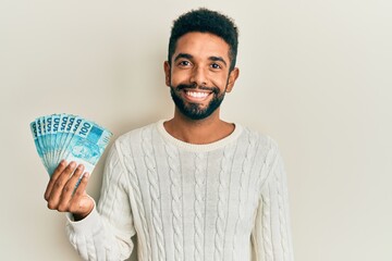 Handsome hispanic man with beard holding 100 brazilian real banknotes looking positive and happy...