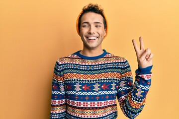 Young handsome man wearing wool hat and colorful sweater smiling with happy face winking at the...
