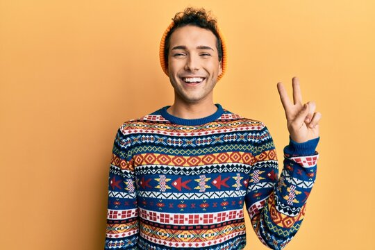 Young handsome man wearing wool hat and colorful sweater smiling with happy face winking at the camera doing victory sign. number two.