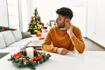 Arab young man sitting on the table by christmas tree smiling with hand over ear listening an hearing to rumor or gossip. deafness concept.