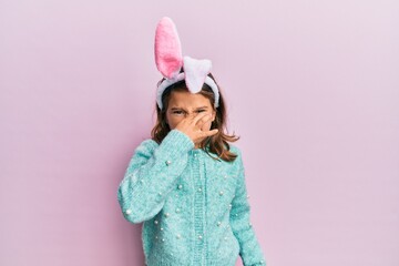 Little beautiful girl wearing cute easter bunny ears smelling something stinky and disgusting, intolerable smell, holding breath with fingers on nose. bad smell