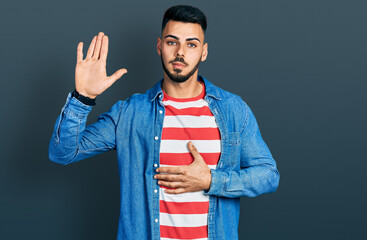 Young hispanic man with beard wearing casual denim jacket swearing with hand on chest and open...