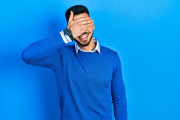 Young hispanic man with beard wearing casual blue sweater smiling and laughing with hand on face covering eyes for surprise. blind concept.