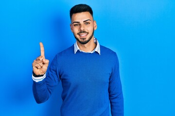Fototapeta premium Young hispanic man with beard wearing casual blue sweater pointing finger up with successful idea. exited and happy. number one.
