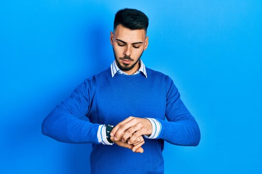 Young hispanic man with beard wearing casual blue sweater checking the time on wrist watch, relaxed and confident
