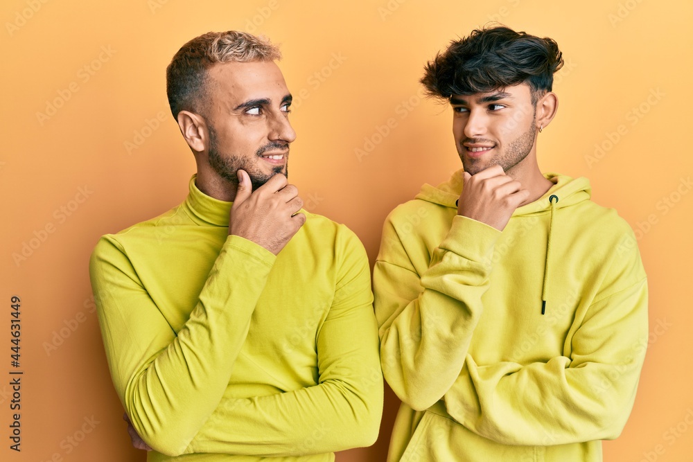 Wall mural Homosexual gay couple standing together wearing yellow clothes with hand on chin thinking about question, pensive expression. smiling with thoughtful face. doubt concept.