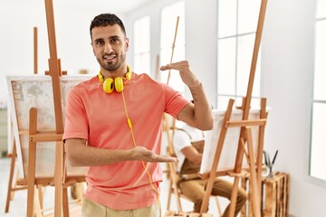 Young hispanic man at art studio gesturing with hands showing big and large size sign, measure symbol. smiling looking at the camera. measuring concept.