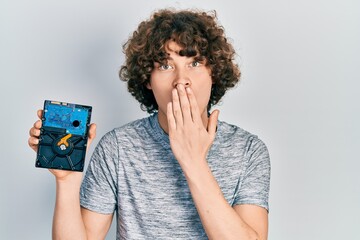 Handsome young man holding hdd disk covering mouth with hand, shocked and afraid for mistake....