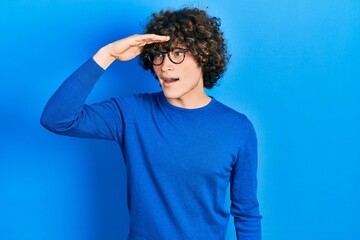 Handsome young man wearing casual clothes and glasses very happy and smiling looking far away with hand over head. searching concept.