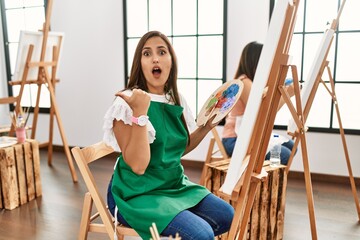 Young hispanic artist women painting on canvas at art studio surprised pointing with hand finger to the side, open mouth amazed expression.