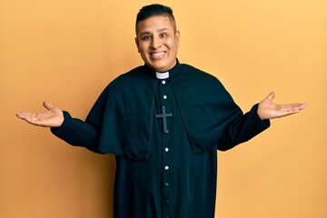 Young latin priest man standing over yellow background smiling showing both hands open palms,...