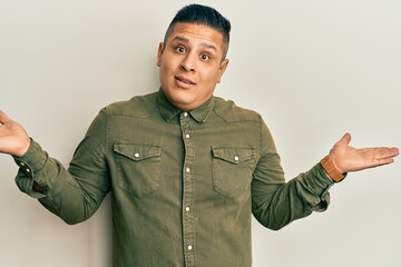 Young latin man wearing casual clothes clueless and confused expression with arms and hands raised....
