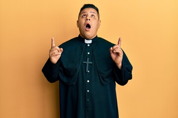 Young latin priest man standing over yellow background amazed and surprised looking up and pointing...
